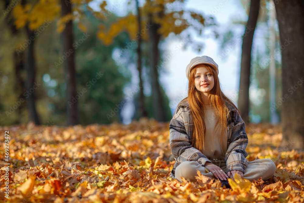 A young pretty girl with long red hair sitting in many falled orange and yellow leaves on a ground in a park. The woman is dressed in a cap and a warm jacket in a cage. Joy, autumn, relax concept. A young pretty girl with long red hair sitting in many falled orange and yellow leaves on a ground in a park. The woman is dressed in a cap and a warm jacket in a cage. Joy, autumn, relax concept.