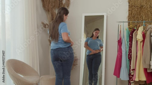 Chubby young adult woman in jeans approaching mirror, touching her abdomen, expressing self-dissatisfaction indoors. Plump lady looking on her reflection, checking fat fold on belly at home