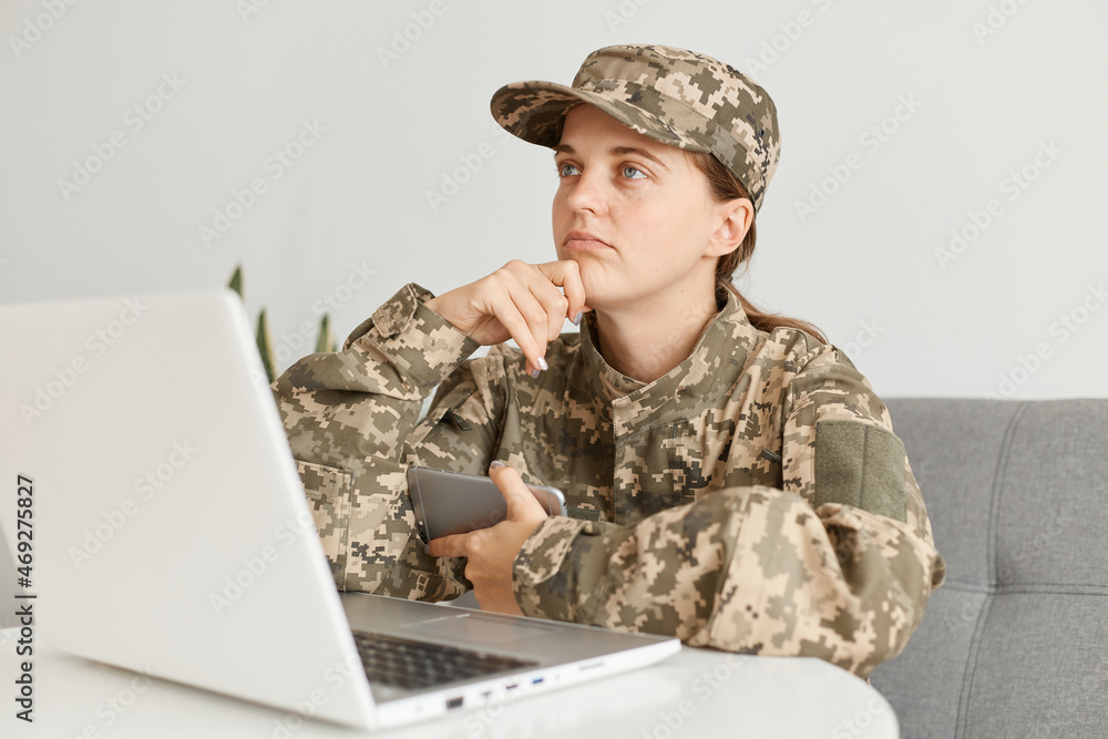 Fototapeta premium Portrait of thoughtful female soldier wearing military costume, posing indoor in light room in front of portable computer with pensive facial expression, looking away.