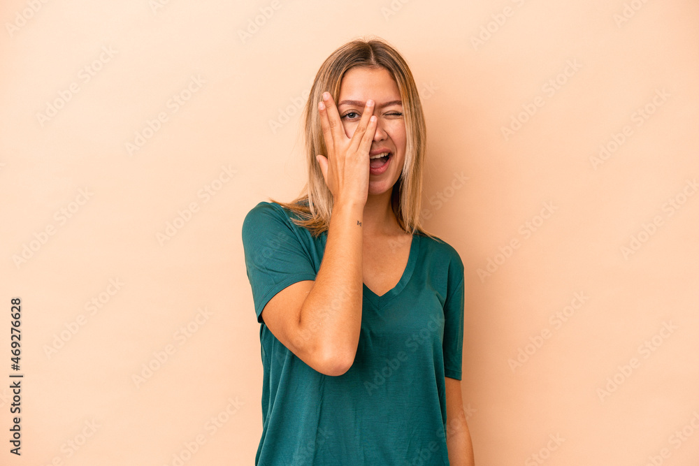 Young caucasian woman isolated on beige background blink at the camera through fingers, embarrassed covering face. Young caucasian woman isolated on beige background blink at the camera through fingers, embarrassed covering face.