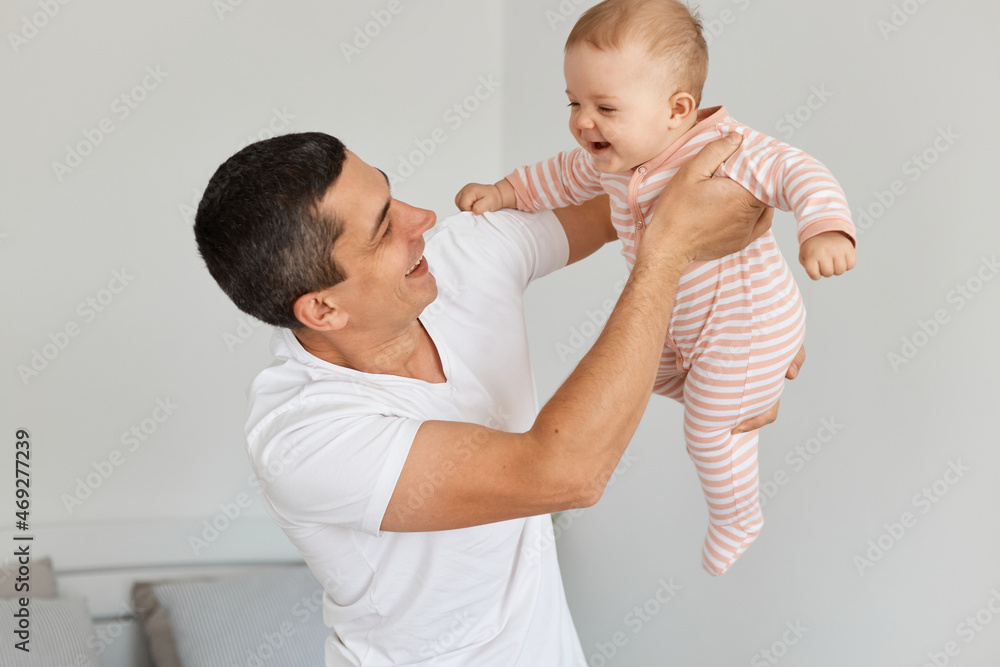 Indoor shot of brunette man wearing white casual t shirt holding his infant daughter, throwing baby in the air and laughing happily, posing at home in light room.