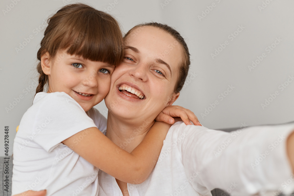 Extremely happy smiling female taking selfie with charming dark haired daughter, looking smiling at cell phone camera and hugging point of view photo, family posing at home.