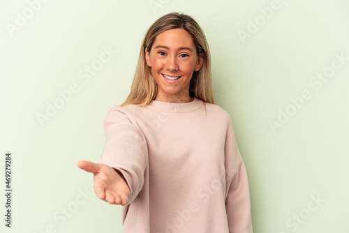 Young caucasian woman isolated on green background stretching hand at camera in greeting gesture.