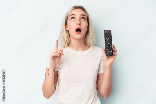 Young caucasian woman holding a tv controller isolated on blue background pointing upside with opened mouth.