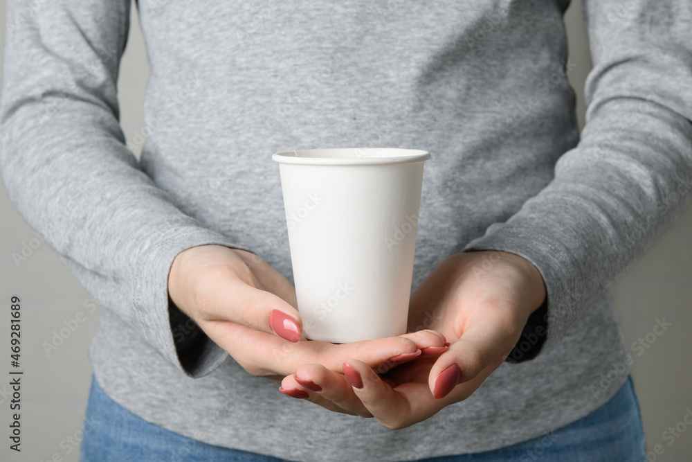 Faceless woman holding blank empty take away paper cup from bottom on neutral grey t-shirt, paper cup mockup with space for presentation in center, side view, close up Faceless woman holding blank empty take away paper cup from bottom on neutral grey t-shirt, paper cup mockup with space for presentation in center, side view, close up