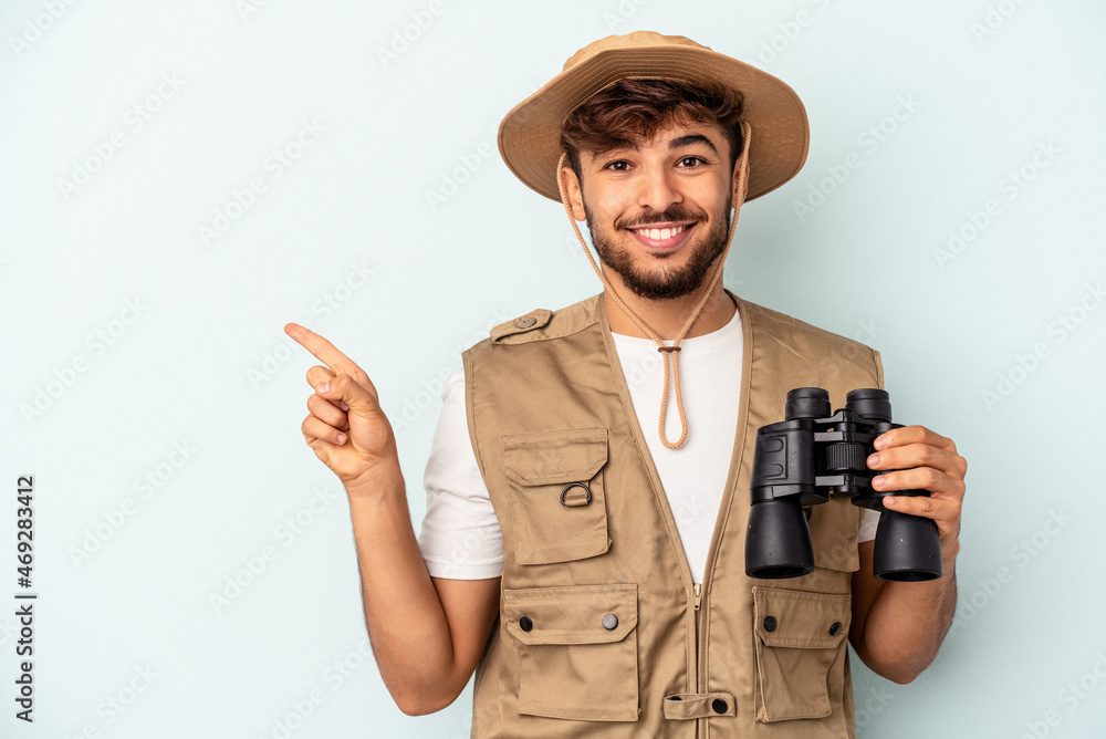 Young mixed race man holding binoculars isolated on blue background smiling and pointing aside, showing something at blank space. Young mixed race man holding binoculars isolated on blue background smiling and pointing aside, showing something at blank space.