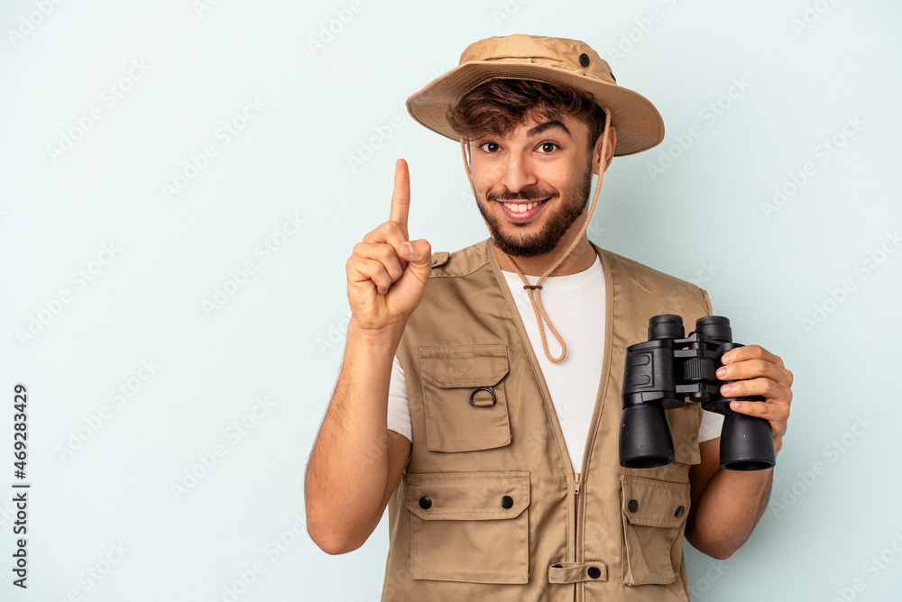 Young mixed race man holding binoculars isolated on blue background showing number one with finger. Young mixed race man holding binoculars isolated on blue background showing number one with finger.