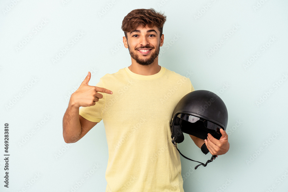 Naklejka premium Young mixed race man holding motorcycle helmet isolated on blue background person pointing by hand to a shirt copy space, proud and confident