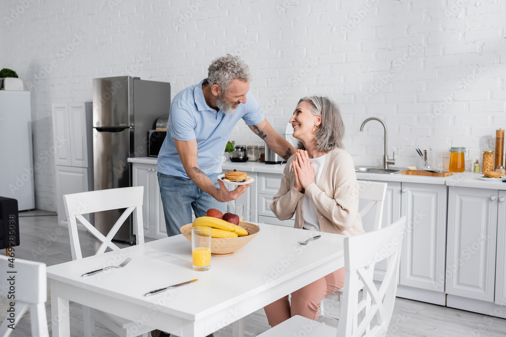 Obraz premium Man holding plate with pancakes while smiling wife showing please gesture in kitchen.