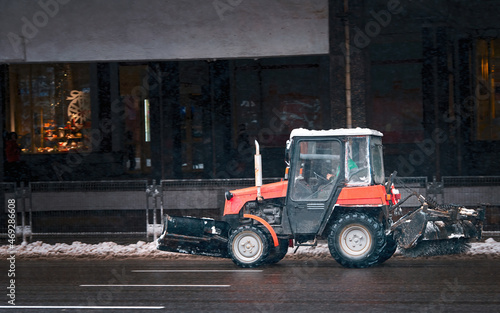 Small tractor with rotary brush and snowplow driving on city road on snowy winter day. Municipal city service cleaning snow , tractor with rotating brush clears road from ice and snow.