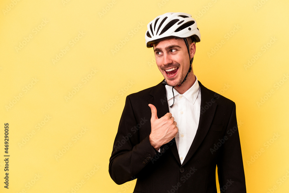 Young caucasian business man wearing a bike helmet isolated on yellow background points with thumb finger away, laughing and carefree. Young caucasian business man wearing a bike helmet isolated on yellow background points with thumb finger away, laughing and carefree.