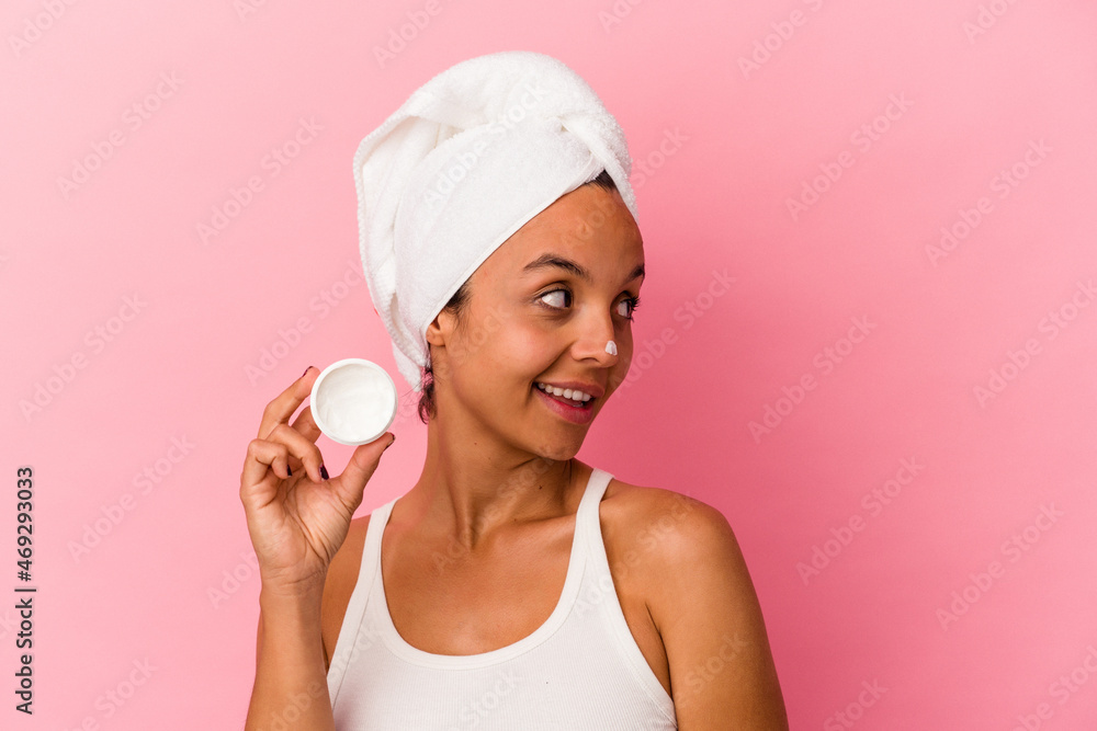 Young mixed race woman holding facial cream isolated on pink background looks aside smiling, cheerful and pleasant. Young mixed race woman holding facial cream isolated on pink background looks aside smiling, cheerful and pleasant.