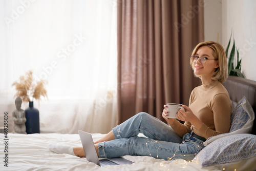 Woman relaxing and drinking cup of hot coffee or tea using laptop computer in the bedroom. Communication and technology concept