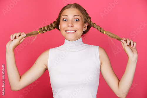 Portrait of cheerful smiling and winking woman holding long hair pigtails on pink background.