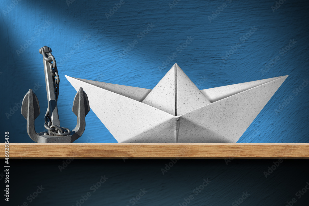Closeup of a white paper boat and a gray metal anchor with chain on a wooden shelf at home, with a blue wall on background.