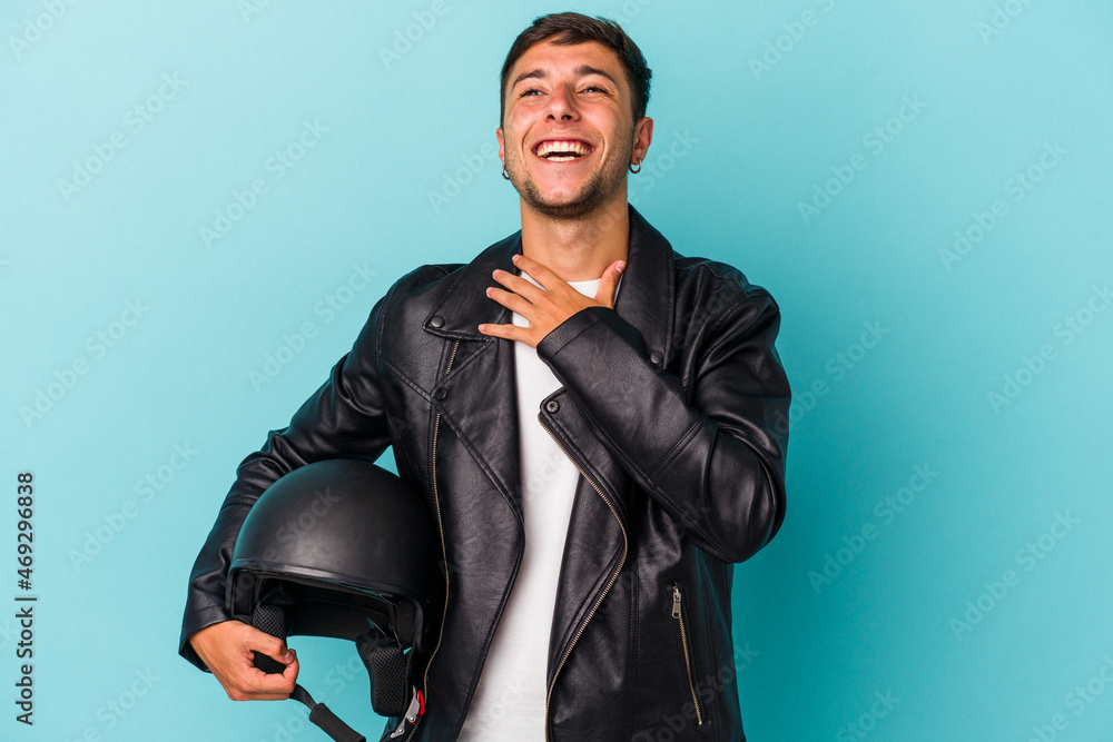 Young biker man holding helmet isolated on blue background laughs out loudly keeping hand on chest. Young biker man holding helmet isolated on blue background laughs out loudly keeping hand on chest.