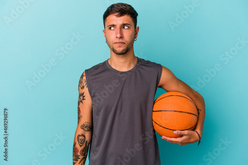 Young caucasian man playing basketball isolated on blue background confused, feels doubtful and unsure.