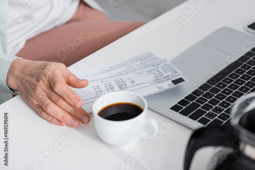 Cropped view of mature woman holding bills near coffee and laptop on table.
