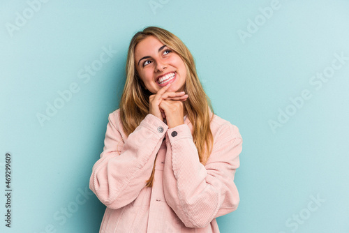 Young caucasian blonde woman isolated on blue background keeps hands under chin, is looking happily aside.