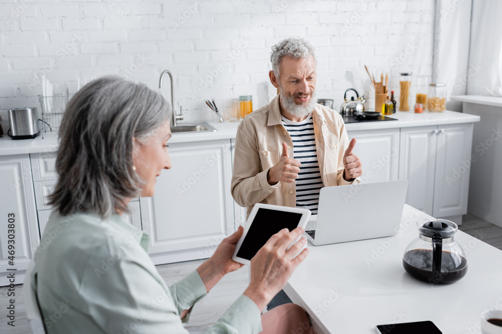Obraz premium Mature man showing thumbs up during video call on laptop near coffee and wife with digital tablet at home.