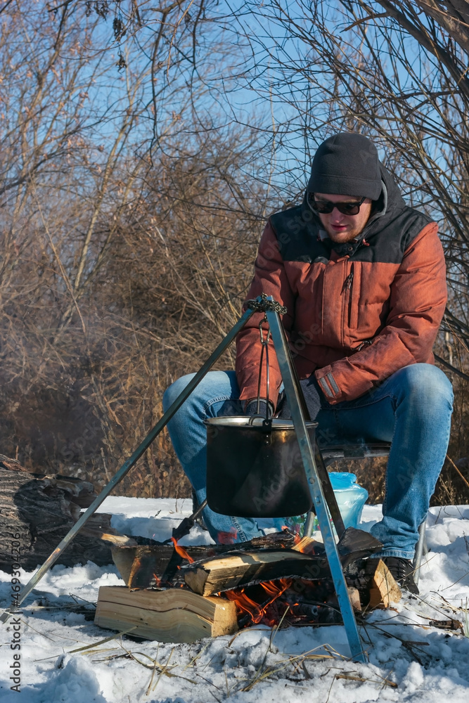 Caucasian man by campfire, pot of soot over bonfire hanging on tripod, winter outdoor cooking at campsite Caucasian man by campfire, pot of soot over bonfire hanging on tripod, winter outdoor cooking at campsite