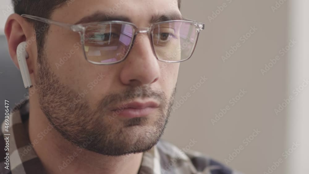 Slowmo close-up of serious bearded Mixed-race programmer in wireless earphones looking at computer screen while running program codes reflecting in his eyeglasses Slowmo close-up of serious bearded Mixed-race programmer in wireless earphones looking at computer screen while running program codes reflecting in his eyeglasses