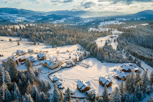 Aerial winter landscape with small village houses between snow covered forest in cold mountains in the evening