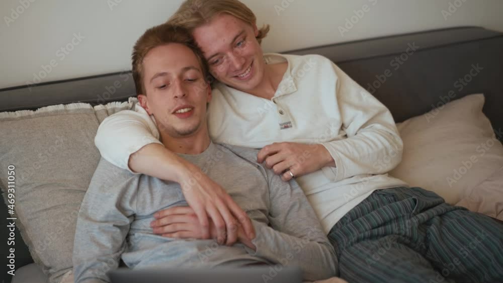 Smiling gay couple looking at laptop and talking while lying on the bed in the room Smiling gay couple looking at laptop and talking while lying on the bed in the room