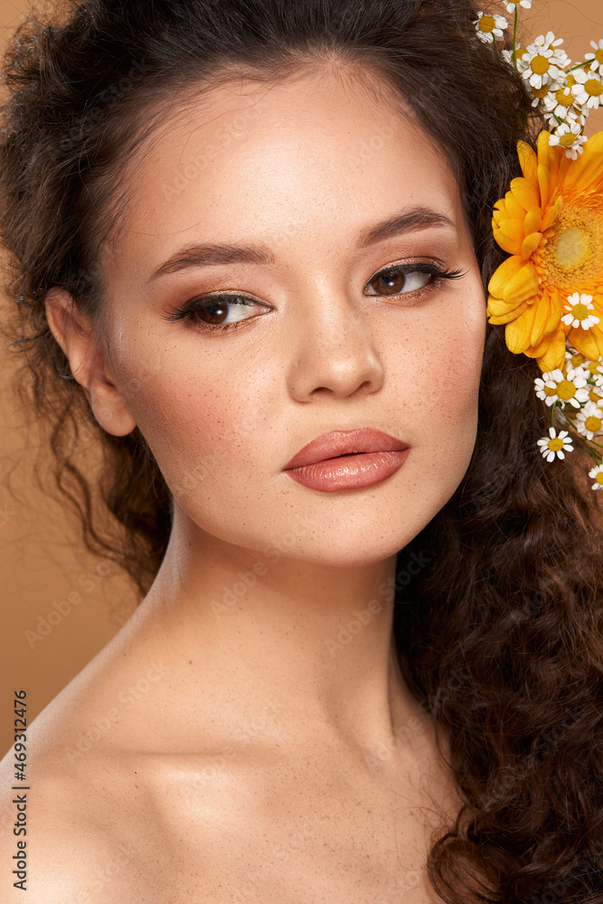 Close-up fashion beauty portrait of fashion female model with flowers in curly hair and perfect make up against beige background