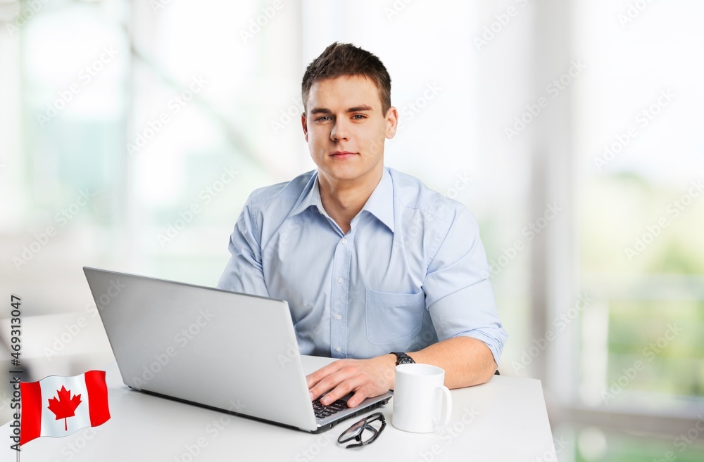 Happy young guy sitting with flag of Canada, using a laptop computer Happy young guy sitting with flag of Canada, using a laptop computer