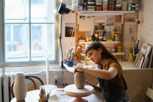 Focused female ceramist shaping potter vase in studio creating ceramics. Artwork for entertainment or small business: artistic workshop owner woman busy with pottery production in creative space room photo