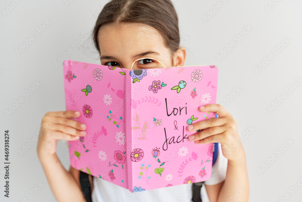 Closeup of a cute pupil covering her face with a colorful book. Schoolgirl smiling during learning from the book. Closeup of a cute pupil covering her face with a colorful book. Schoolgirl smiling during learning from the book.