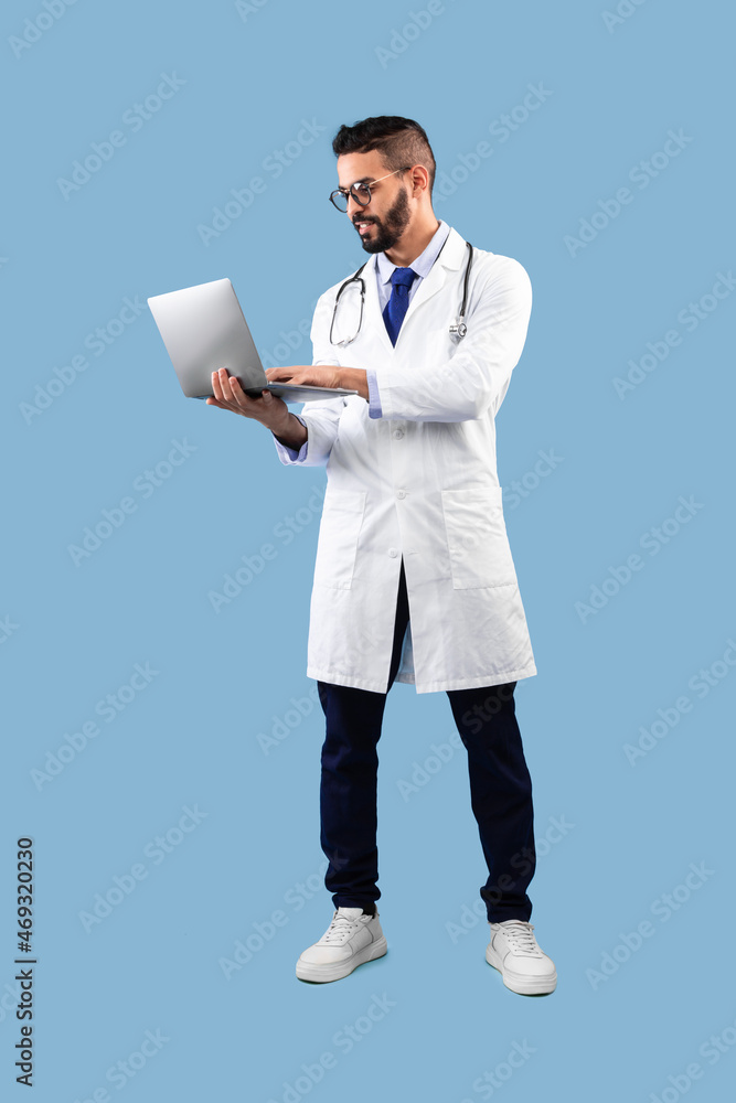 Arabic Male Doctor Using Laptop Standing Over Blue Background, Vertical Arabic Male Doctor Using Laptop Standing Over Blue Background, Vertical