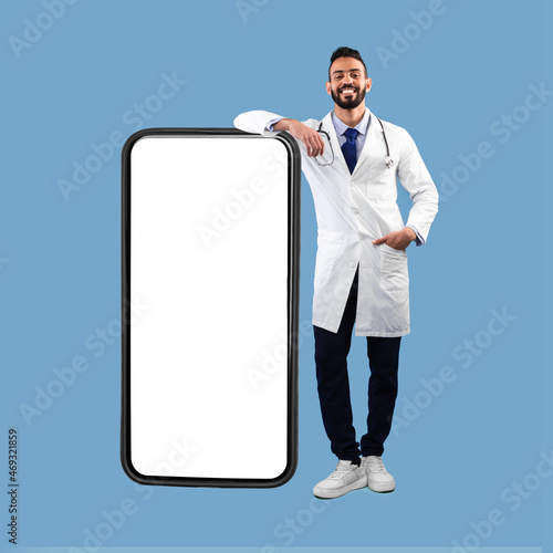 Middle Eastern Male Doctor Leaning On Big Smartphone, Blue Background, Square