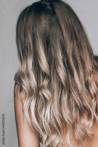 Close-up of the wavy blonde hair of a young blonde woman isolated on a gray background. Result of coloring, highlighting, perming. Beauty and fashion. Rear view. Vertical shot