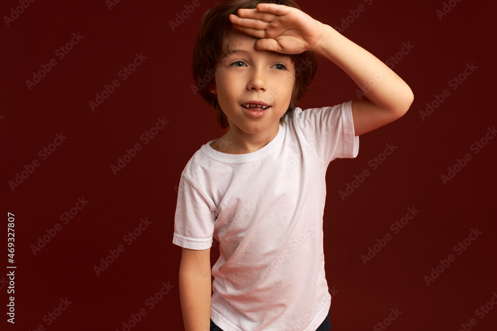 Portrait of tired boy, holding hand over his forehead, smiling, dressed in white, looking aside with happy, joy face, isolated on red studio wall with copy space for your advertising content Portrait of tired boy, holding hand over his forehead, smiling, dressed in white, looking aside with happy, joy face, isolated on red studio wall with copy space for your advertising content