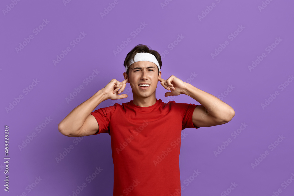 Annoyed young sportsman or coach plugs ears with fingers, suffering from loud noise or music, promotes healthy lifestyle, wears red t-shirt and white headband, isolated on violet background. Annoyed young sportsman or coach plugs ears with fingers, suffering from loud noise or music, promotes healthy lifestyle, wears red t-shirt and white headband, isolated on violet background.