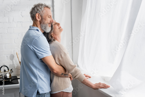 Middle aged man embracing wife near window in kitchen.