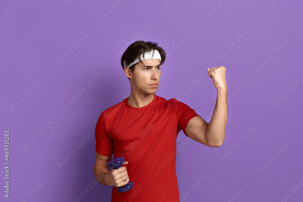 Serious sportsman is holding dumbbell in hand and showing fist, ready to fight for limited offer, promotes healthy lifestyle, wears red t-shirt and white headband, isolated on violet background. Serious sportsman is holding dumbbell in hand and showing fist, ready to fight for limited offer, promotes healthy lifestyle, wears red t-shirt and white headband, isolated on violet background.