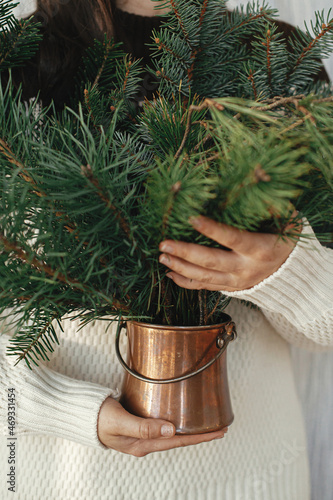 Woman holding pine and fir branches in vintage vase in rustic room. Preparations for winter holidays