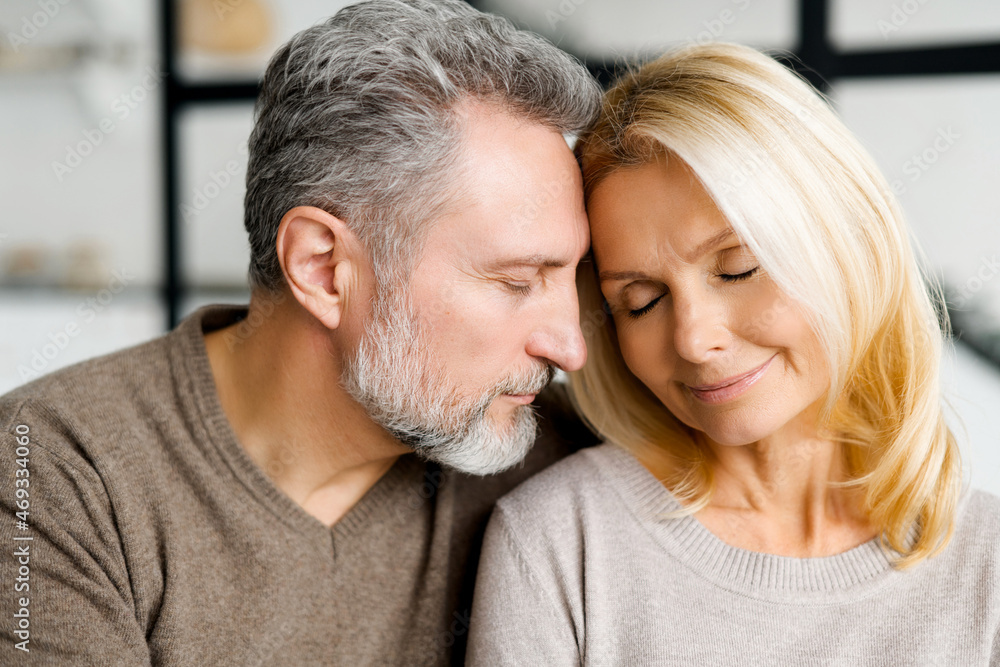 Gentle portrait of romantic serene middle-aged couple in embraces at home. Mature spouses in love sitting on the sofa with eyes closed, feels appeasement and calm. Bonding and love concept Gentle portrait of romantic serene middle-aged couple in embraces at home. Mature spouses in love sitting on the sofa with eyes closed, feels appeasement and calm. Bonding and love concept