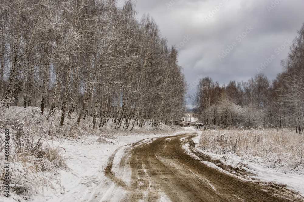 Autumn landscape with the first snowfall in the Urals (Russia). Birches with loose leaves artistically bent to the road leading to the village. Wet tire tracks stand out in relief against white snow Autumn landscape with the first snowfall in the Urals (Russia). Birches with loose leaves artistically bent to the road leading to the village. Wet tire tracks stand out in relief against white snow