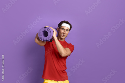 Smiling handsome athlete is carrying heavy fitness or yoga mat on shoulder from gym, holding it with both hands, wears red t-shirt, yellow shorts and white headband, isolated on violet background.