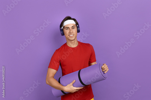 Young smiling athlete or coach is holding fitness or yoga mat with hands, leaving sport club after training, wearing red t-shirt, blue headphones and white headband, isolated on violet background.