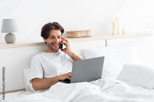 Cheerful attractive millennial european man works on laptop, speaks on phone with client on bed
