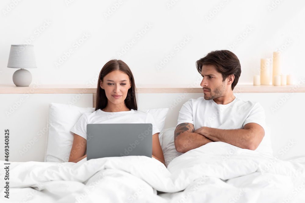 Disgruntled upset caucasian young husband looking at smiling woman chatting on social media Disgruntled upset caucasian young husband looking at smiling woman chatting on social media
