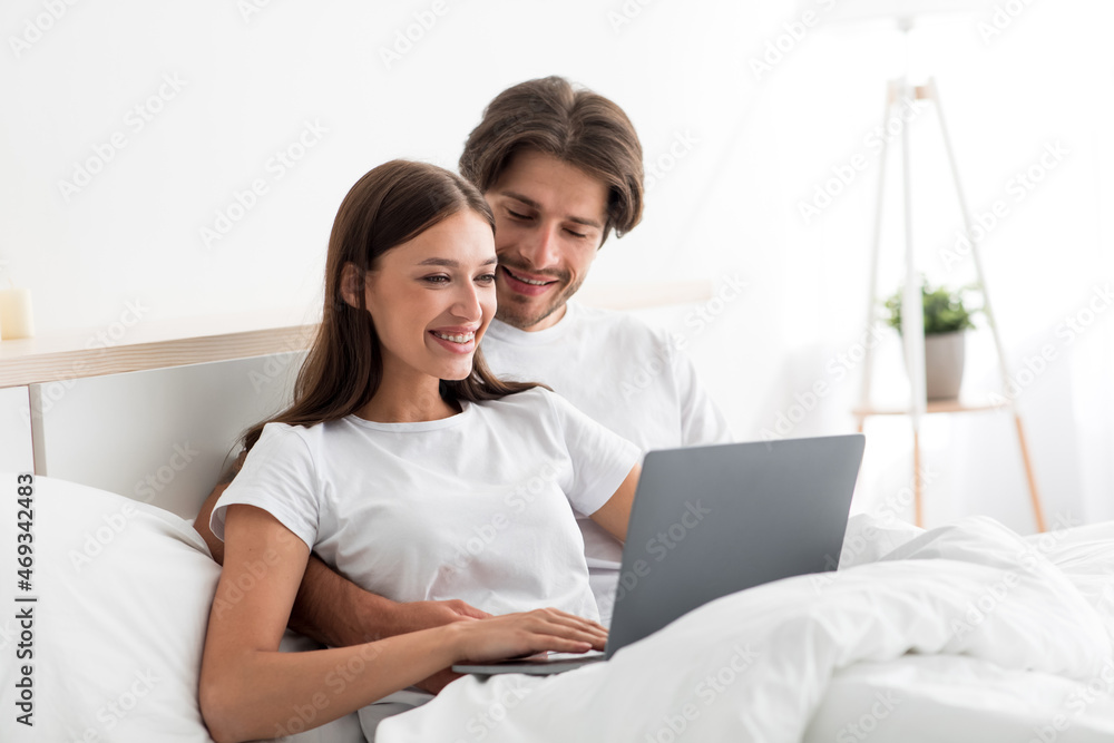 Happy pretty millennial caucasian lady and guy look at laptop chatting or making video call on bed Happy pretty millennial caucasian lady and guy look at laptop chatting or making video call on bed