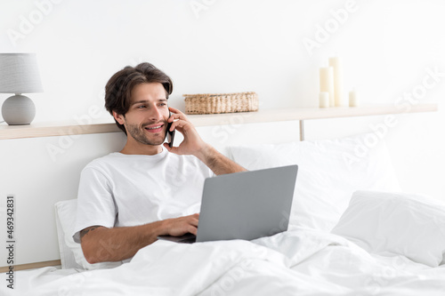Smiling handsome millennial caucasian guy works on laptop, speaks by phone with client