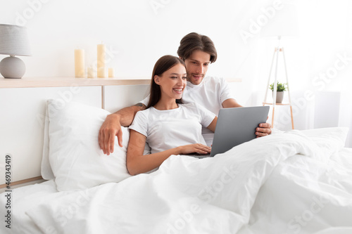 Smiling caucasian young husband and wife hugging, sitting on bed and watch video at laptop