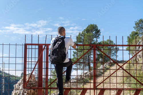 Young man with backpack climbing through a gate with metal fence. 